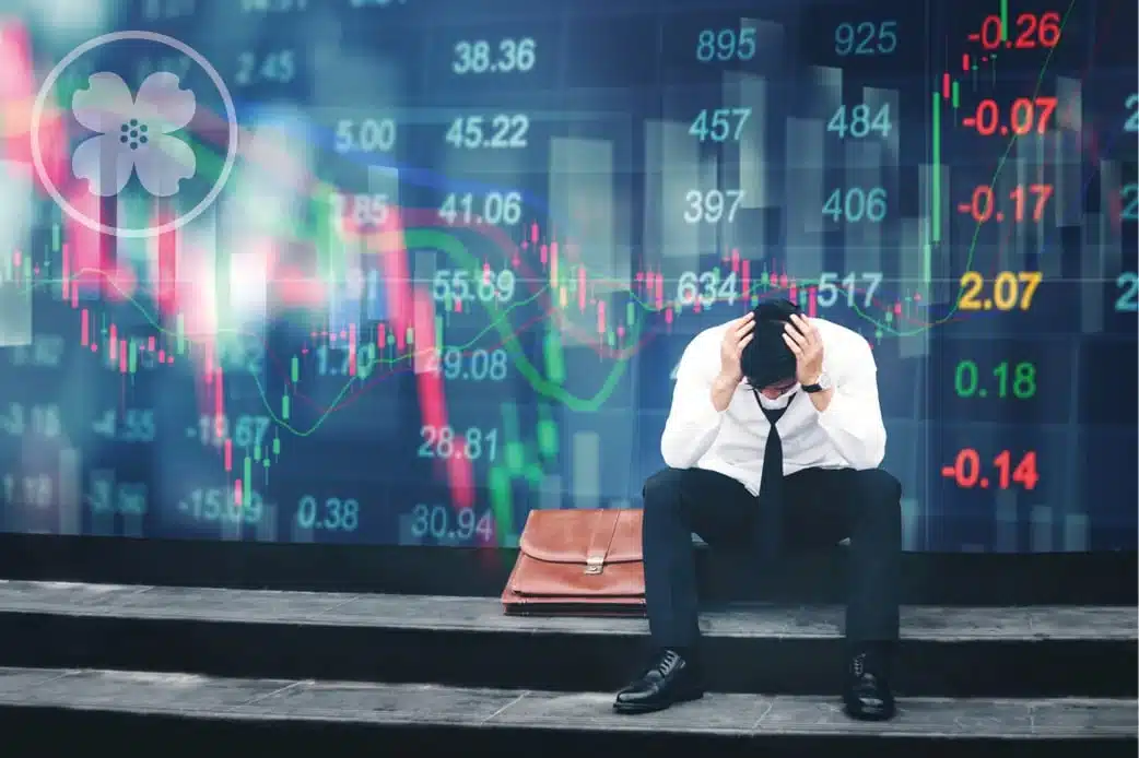 Man sitting on the ground looking down with his hands over his head stressed about the stock market.